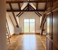 An empty attic space with exposed wooden beams, a large window with decorative ironwork, and a patterned wooden floor.