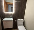 Modern bathroom with a white sink, chrome faucet, toilet, and illuminated mirror above the sink. The walls are tiled in dark gray, and the floor is tiled in beige.