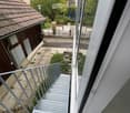 Balcony with metal stairs leading down to a small outdoor area with concrete flooring, greenery, and a wooden shed.