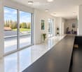 Modern, minimalist hallway with large glass windows, polished marble floors, recessed lighting, and a sleek black table with a stool. Potted plants and artworks adorn the walls.