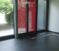 Empty room with dark tiled floor, large sliding glass doors with red blinds, and a view of greenery outside.