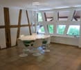 Modern dining room with a white round table, six white chairs, pendant lights, and large windows with wooden shutters.