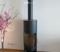 Modern black wood-burning stove with a tall chimney, placed on a glass platform in a room with wooden flooring and white walls. A vase with pink flowers is visible to the left.