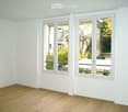 Empty room with wooden flooring and large windows, showing a view of the outside area with trees and a building.
