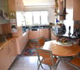 Bright kitchen with light wooden cabinets, a round wooden dining table with four chairs, a sink, and a stove. The room features a colorful rug, framed pictures, and various kitchen items on the counters.