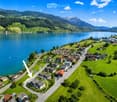 Aerial view of a small village nestled by a lake, surrounded by green hills and mountains. The village features numerous houses with varied architectural styles, roads, and a calm lake reflecting the surrounding landscape.