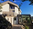 Modern two-story house with white exterior, large windows, balcony, and glass-enclosed patio area with dining table and chairs.