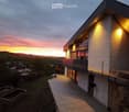 Modern two-story house with a balcony, illuminated exterior lighting, and a scenic sunset view over a rural landscape.