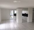 Empty room with white tiled flooring, white walls, and large windows. The space includes a small kitchen area with white cabinets and a countertop.