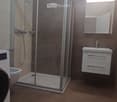 Modern bathroom with a glass-enclosed shower, a white sink with a mirrored cabinet, and a washing machine. The walls are tiled in neutral tones, and the floor is covered with brown tiles.
