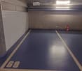 Underground parking space with blue floor, marked parking spots, and a single traffic cone.