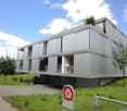 Modern, multi-story building with a sleek, grey concrete facade and large windows, elevated on stilts with a landscaped lawn and trees surrounding it.