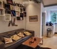Modern living room with a brown leather sofa, wooden coffee table, bookshelves, and a small kitchen area visible through an open doorway.