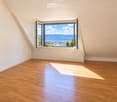 Empty room with wooden flooring and white walls, featuring a large window offering a scenic view of a lake and mountains.