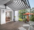 Wooden deck with white wicker chairs, small table, outdoor umbrella, and a large ceramic vase. The deck is covered by a black and white striped awning, with a view of a garden and trees.