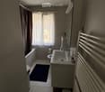 Modern bathroom with a sink, toilet, bathtub, radiator, and a small stool near the sink. The room features a window with sheer curtains and a dark bathmat on the floor.