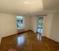 Empty room with wooden herringbone flooring, white walls, large windows with white curtains, radiator, and a ceiling light fixture.