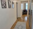 A well-lit hallway with wooden flooring, a patterned rug, and three framed black-and-white pictures on the wall. The hallway leads to a living room area with a sofa and side table visible in the background.
