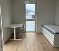 A small, modern room with light wooden flooring, a white desk with a chair, and a built-in bed with a mattress and storage unit.