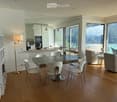 Open-plan living and dining area with modern furniture, large windows overlooking a scenic lake, and a kitchen area with cabinets and appliances.