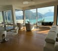 Modern living room with large windows offering a scenic mountain and lake view. Features a sectional sofa, coffee table, dining table with chairs, and several lamps.