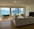 Modern living room with large windows offering a scenic mountain and lake view, featuring a grey sectional sofa, coffee table, armchairs, ottoman, and a television.