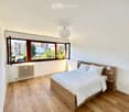 Bright bedroom with wooden flooring, large windows, a neatly made bed with white linens, and a nightstand with a lamp.