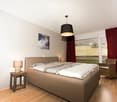 Modern bedroom with a large bed, nightstands, lamps, a wicker chair, and artwork on the walls. Large windows with red curtains offer a view of the countryside.