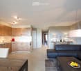 Modern open-plan living and dining area with a sleek black sectional sofa, a wooden coffee table with fruit, a dining table with chairs, and a contemporary kitchen featuring light wood cabinets, stainless steel appliances, and a central island.