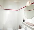 Modern bathroom with white tiles, a toilet, sink, mirror, and towel. Features a red accent border on the wall and a small trash can near the toilet.