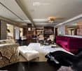 Elegant living room with patterned wallpaper, dark hardwood floors, and a mix of plush seating including a red velvet sofa, floral-patterned armchairs, and a white ottoman. A coffee table sits in the center, flanked by additional seating and a large mirro