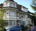 Two-story house with white exterior, multiple windows, and a balcony on the upper floor. A blue car is parked in front of the house.