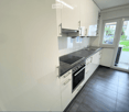 Modern kitchen with white cabinets, dark countertops, induction cooktop, oven, and sink. Large window offers a view of the garden.