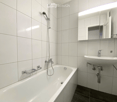 Modern bathroom with white tiled walls and floor, featuring a bathtub with a handheld shower, a sink with a modern faucet, and a large mirror with built-in lighting.