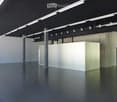 Empty, unfurnished room with a polished concrete floor, white walls, and a single white partition. Exposed metal beams and industrial lighting fixtures are visible on the ceiling.