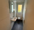 Compact bathroom with a white sink, chrome faucet, toilet, and a small mirror above the sink. The room has light-colored tiles and a dark floor.