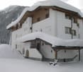 A large, two-story white house with wooden accents, snow-covered roof and surrounding area, set against a mountainous backdrop.