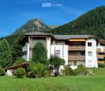 A multi-story white building with balconies and wooden railings, surrounded by greenery and mountains.