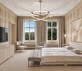 Elegant bedroom with a large bed, bench at the foot, two armchairs, dresser, television, and artwork on the wall. The room features large windows with curtains and a modern chandelier.