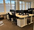 Modern office space with large windows, desks, chairs, multiple computer monitors, keyboards, mice, and a server rack. A large exercise ball and trash cans are also visible.