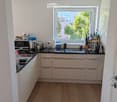 Modern kitchen with light-colored cabinets, wooden flooring, and various appliances including a microwave, toaster, coffee maker, and knife block. A window offers a view of greenery outside.
