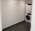 A small, modern laundry room with a black tiled floor, white walls, and a white washing machine positioned against the wall.