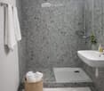 A bathroom featuring a modern, stylish design with tiled floors and walls. A shower stall with a glass door and silver shower head is present. There is a white sink with a faucet and a mirror on the wall. Two white towels are on a towel rack and a wicker