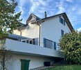 Modern two-story house with white exterior, blue shutters, and a large balcony on the first floor.