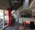 Open-plan living and dining area with a dining table, chairs, kitchen area, and potted plants. Features a large window with a red curtain and a view of a balcony with outdoor furniture.
