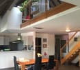 Open-plan living and dining area with a wooden dining table, black chairs, modern kitchen, staircase leading to an upper level, and decorative elements like a flower vase and picture frames.