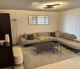 Modern living room with a grey sectional sofa, coffee table, ottoman, and a floor lamp. A large mirror hangs on the wall, and a small table holds decorative items.