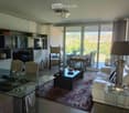 Elegant living room with large windows offering a scenic view, featuring a cream-colored sofa, glass coffee table, glass dining table with chairs, and a patterned area rug.