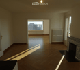 Empty room with wooden flooring, large windows, and a decorative fireplace. Natural light streams in from the windows.
