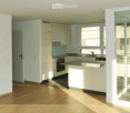 Modern kitchen with white cabinets, a central island, and a dark countertop. Features an oven, sink, and faucet, with large windows allowing natural light to fill the space.